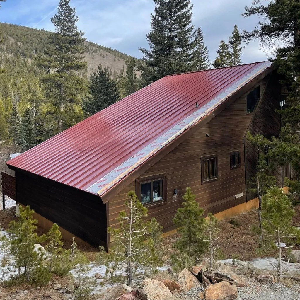 Red metal roof panels partially installed on a wood-sided mountain home, with roofing underlayment visible along the eave